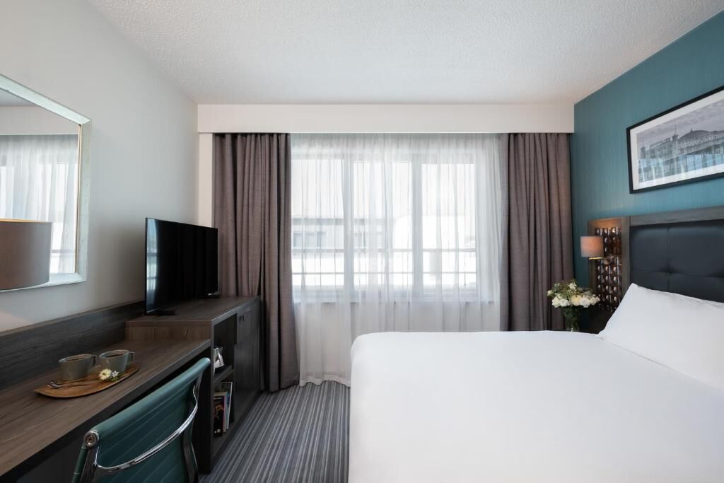 Une chambre d’hôtel avec un lit, un bureau avec une télévision, une fenêtre avec des rideaux et un tableau encadré au mur.