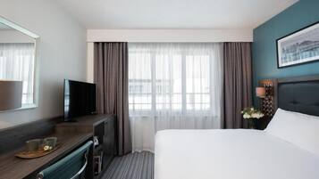 Une chambre d’hôtel avec un lit, un bureau avec une télévision, une fenêtre avec des rideaux et un tableau encadré au mur.