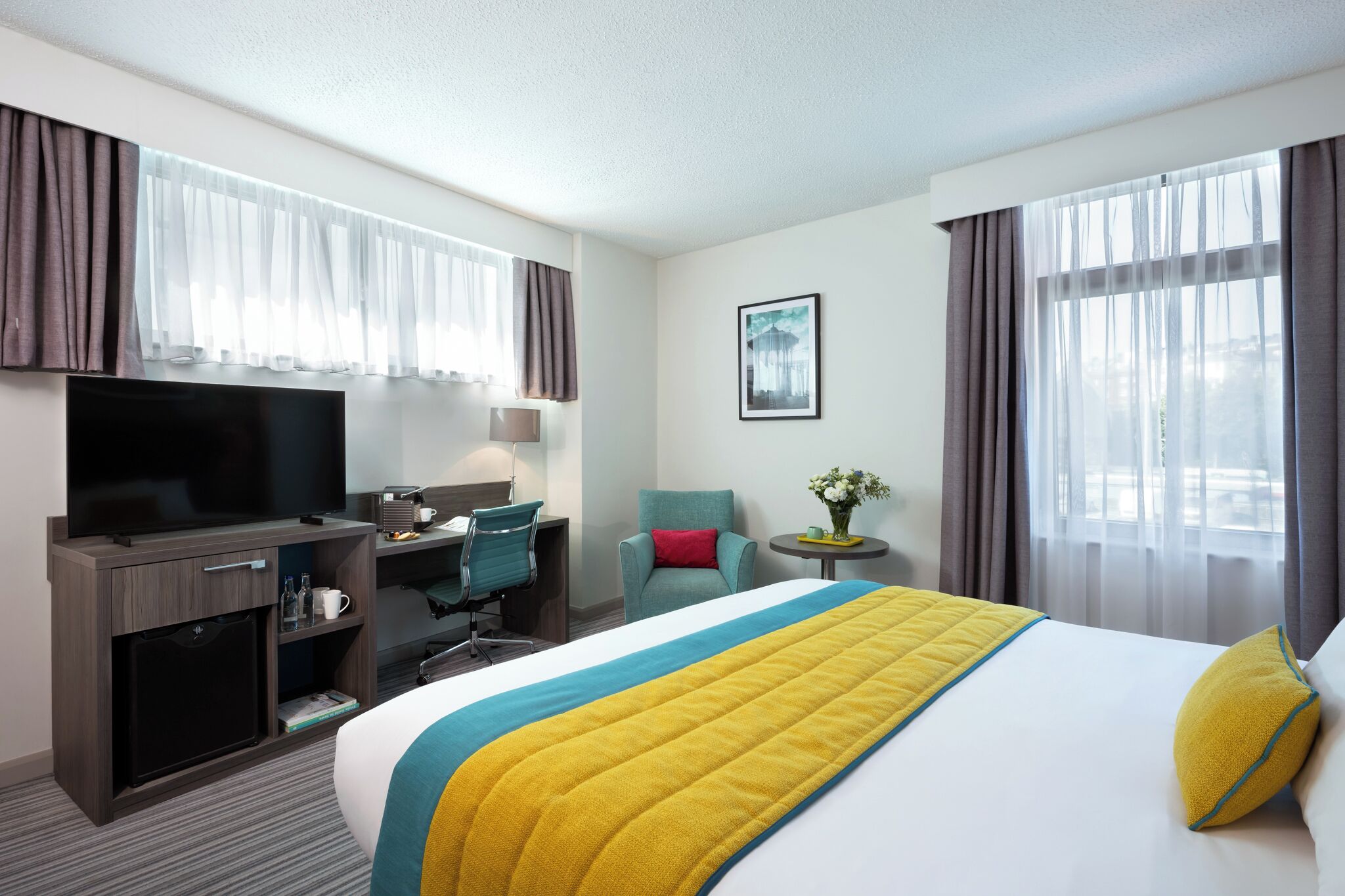 A hotel room with a bed, a TV, a desk, a chair, and a small table with a vase.