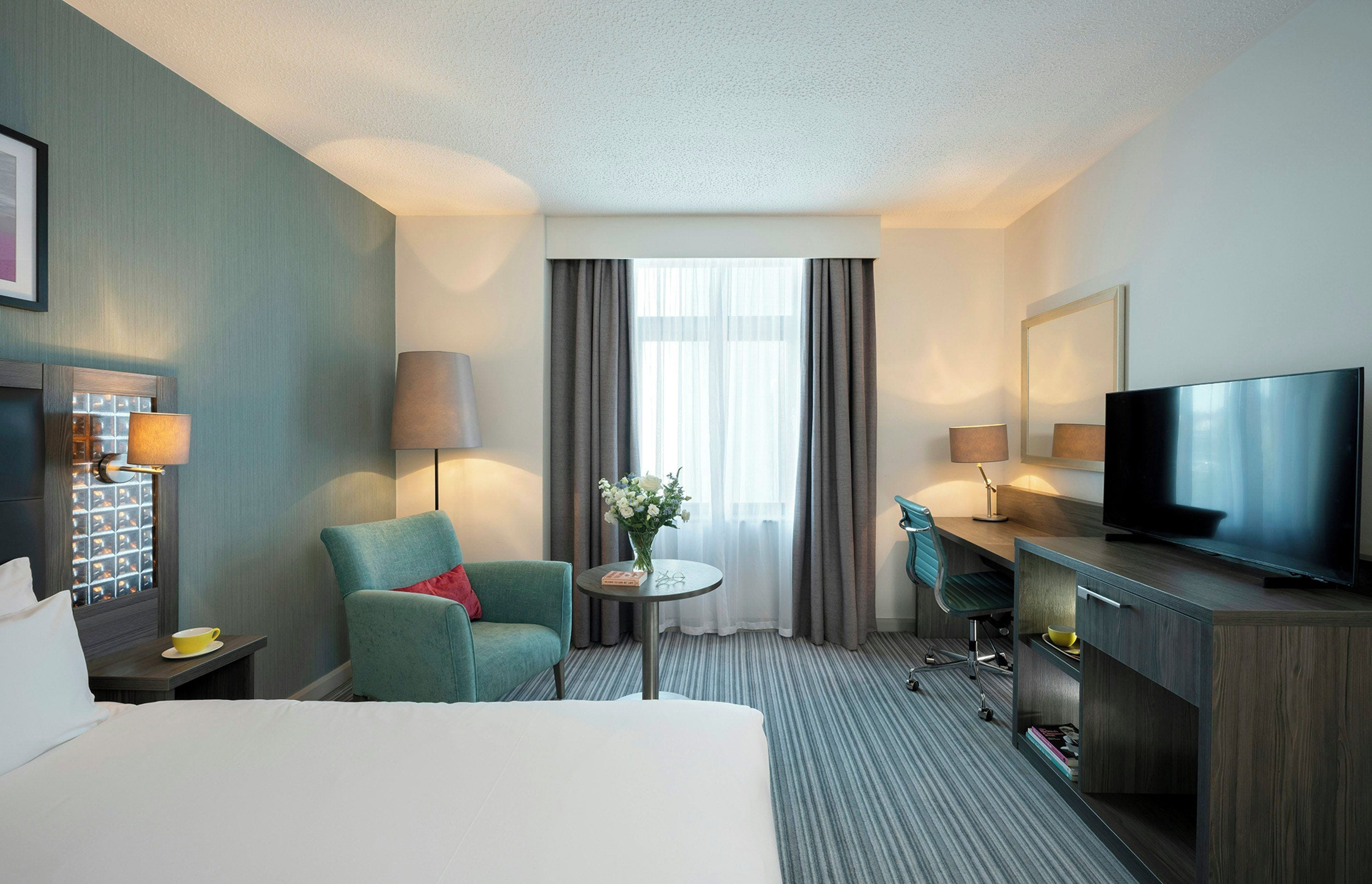 A hotel room with a bed, a desk with a TV, a chair, a small table with a vase, and a window with curtains.