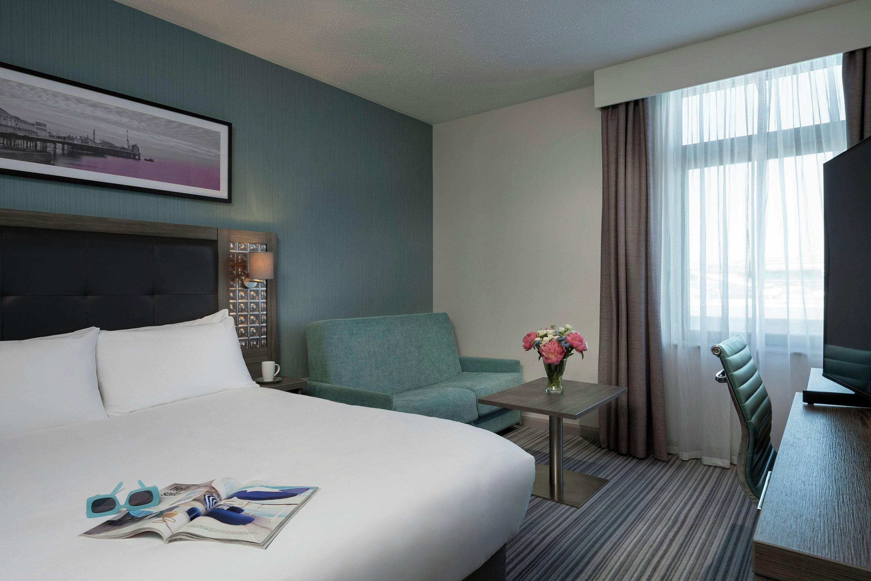 A hotel room with a bed, a seating area with a chair and a table, a window with curtains, and a wall-mounted TV.