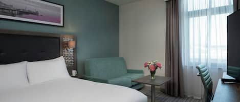 A hotel room with a bed, a seating area with a chair and a table, a window with curtains, and a wall-mounted TV.
