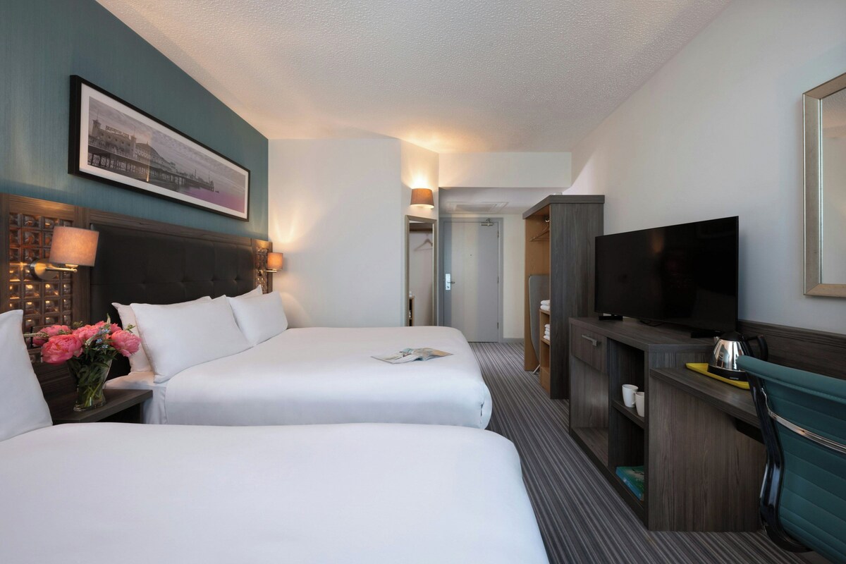 A hotel room with two beds, a television, a desk with a lamp, and a framed picture on the wall.