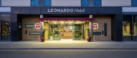 Exterior del Hotel Leonardo con fachada moderna, amplio acceso de vidrio y plantas en macetas a la entrada.