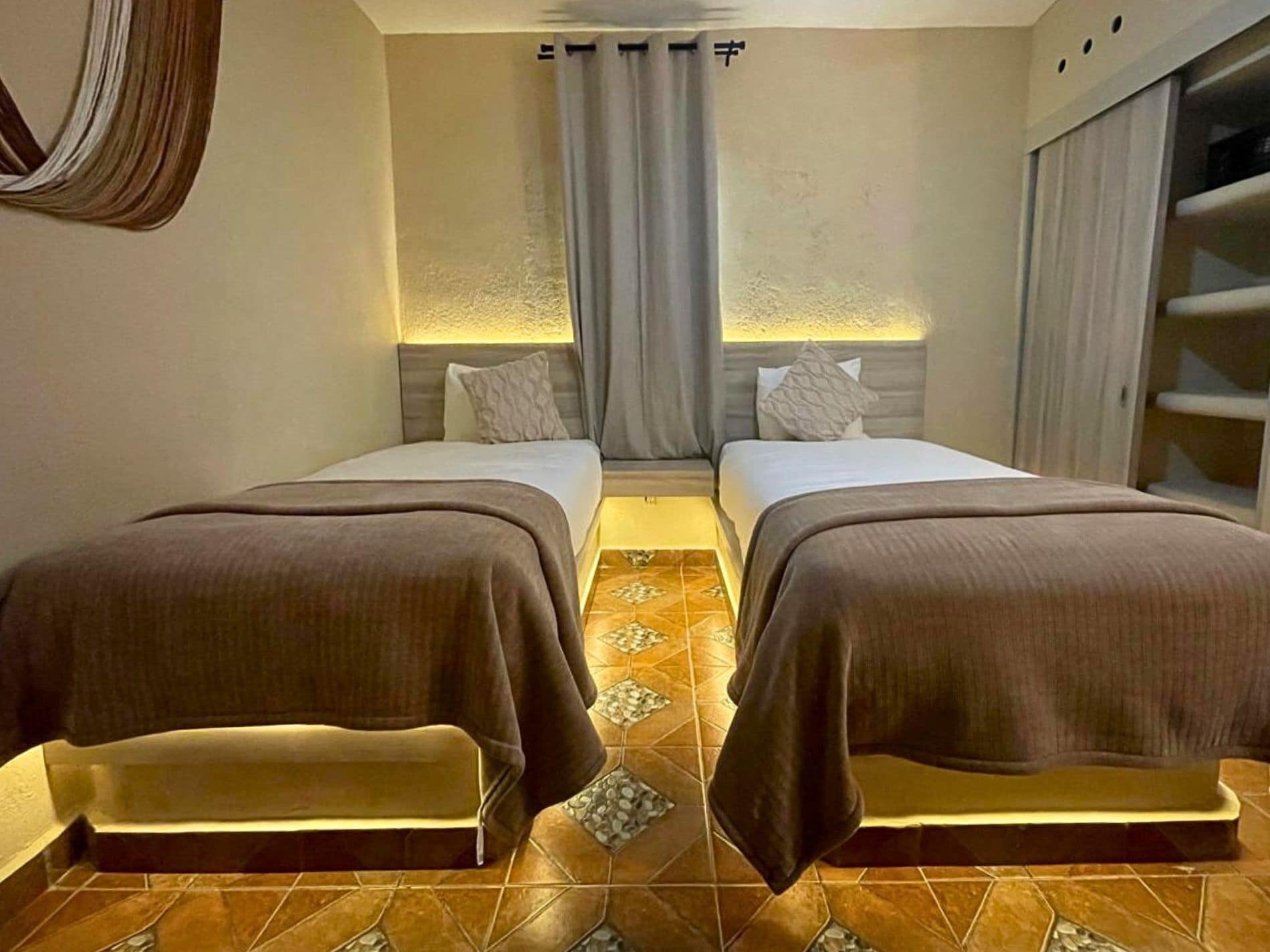 A hotel room with two beds, a bench, a large window with curtains, and a tiled floor.