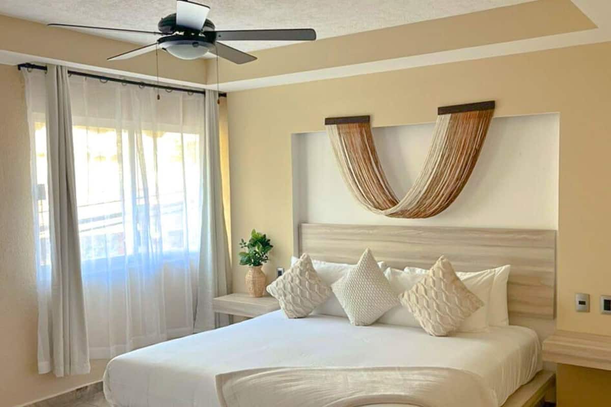 A bedroom with a bed, curtains, a ceiling fan, a wall-mounted plant, and a decorative wall piece.