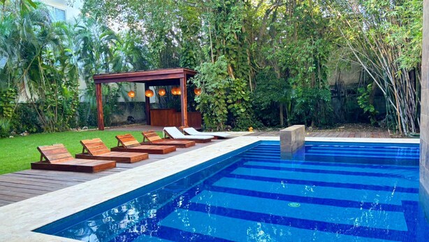 Una piscina rectangular con azulejos azules y una zona de terraza de madera con sillones de descanso.