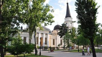 A large building with classical architecture, including columns and a dome, surrounded by a park with trees and a paved walkway.