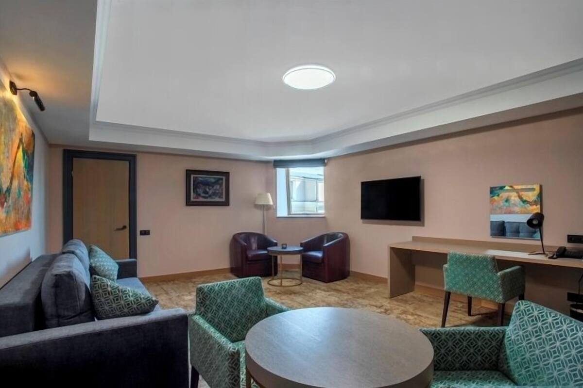Et moderne hotelværelse med sofa, to lænestole, et rundt bord, et fjernsyn og kunst på væggene.