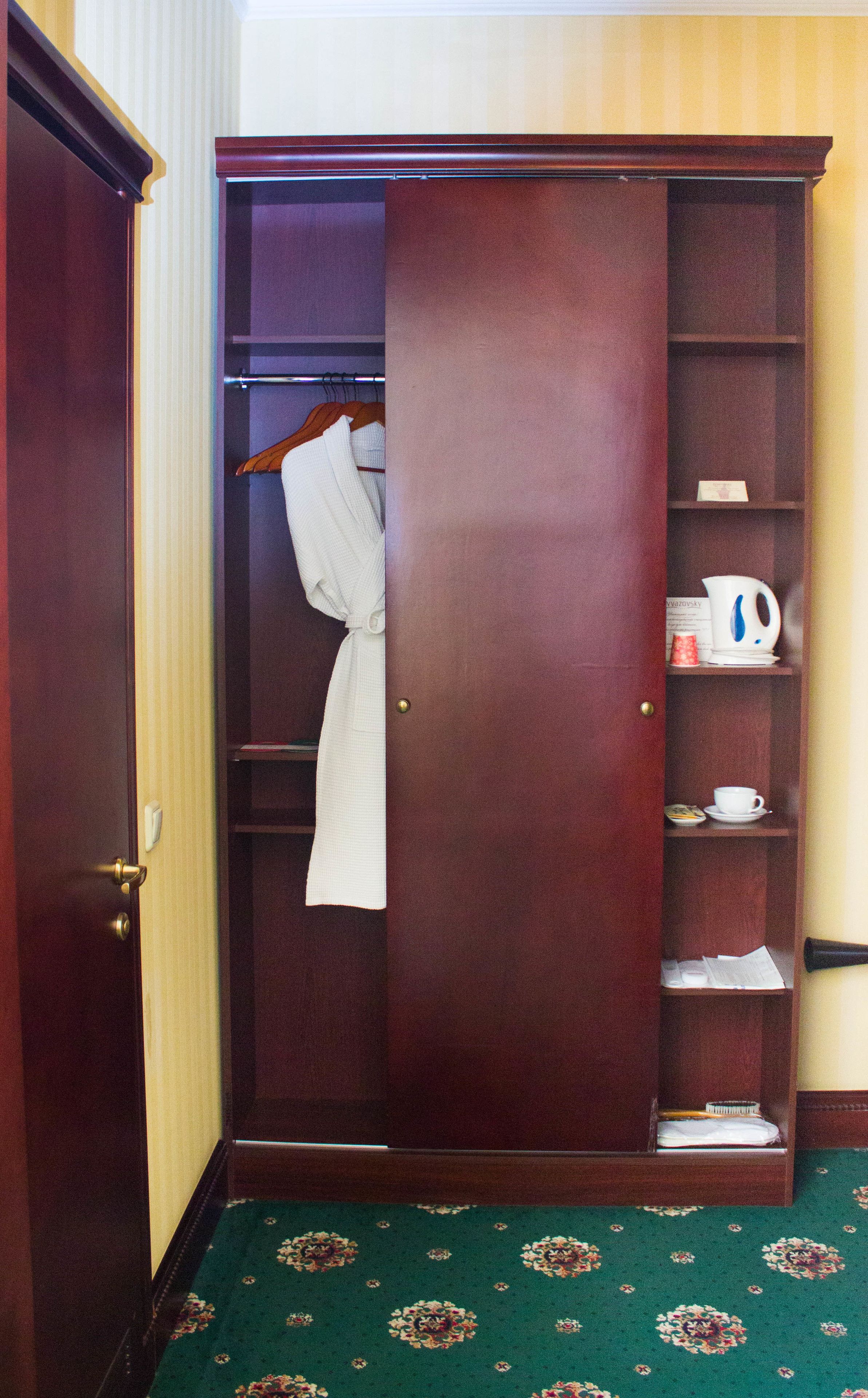 A wooden wardrobe with a robe hanging on the door, a shelf with a kettle, cups, and a small book.