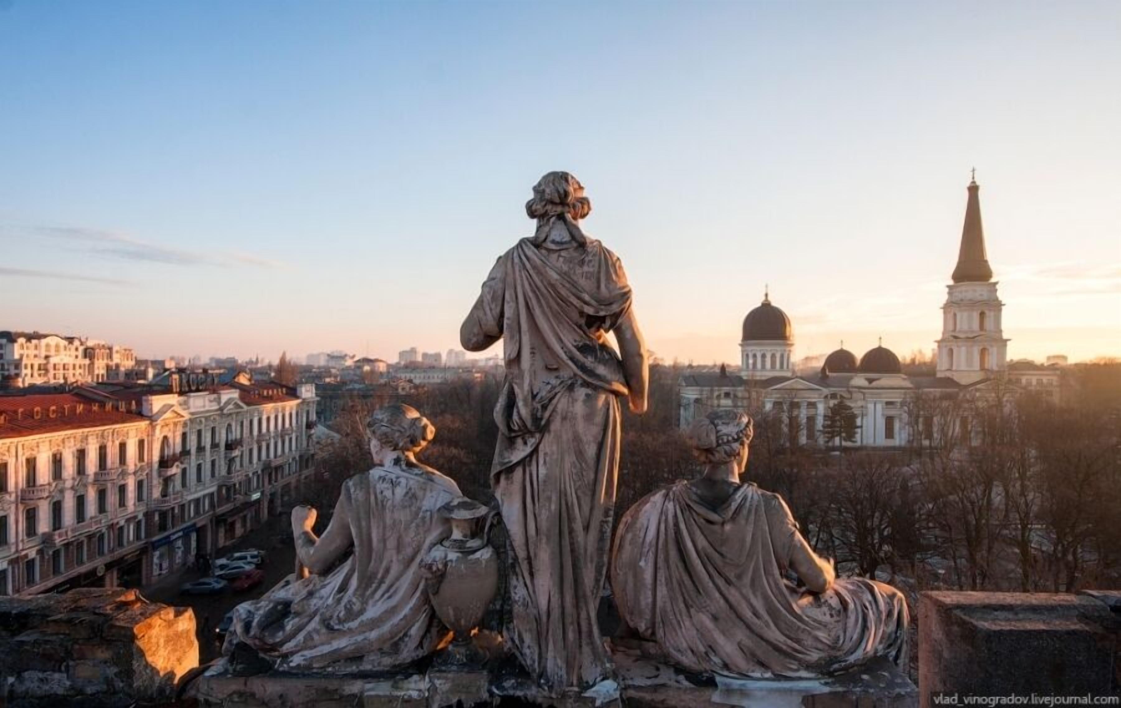 A statue of a seated figure with a draped robe, overlooking a cityscape with historical buildings and a church spire.