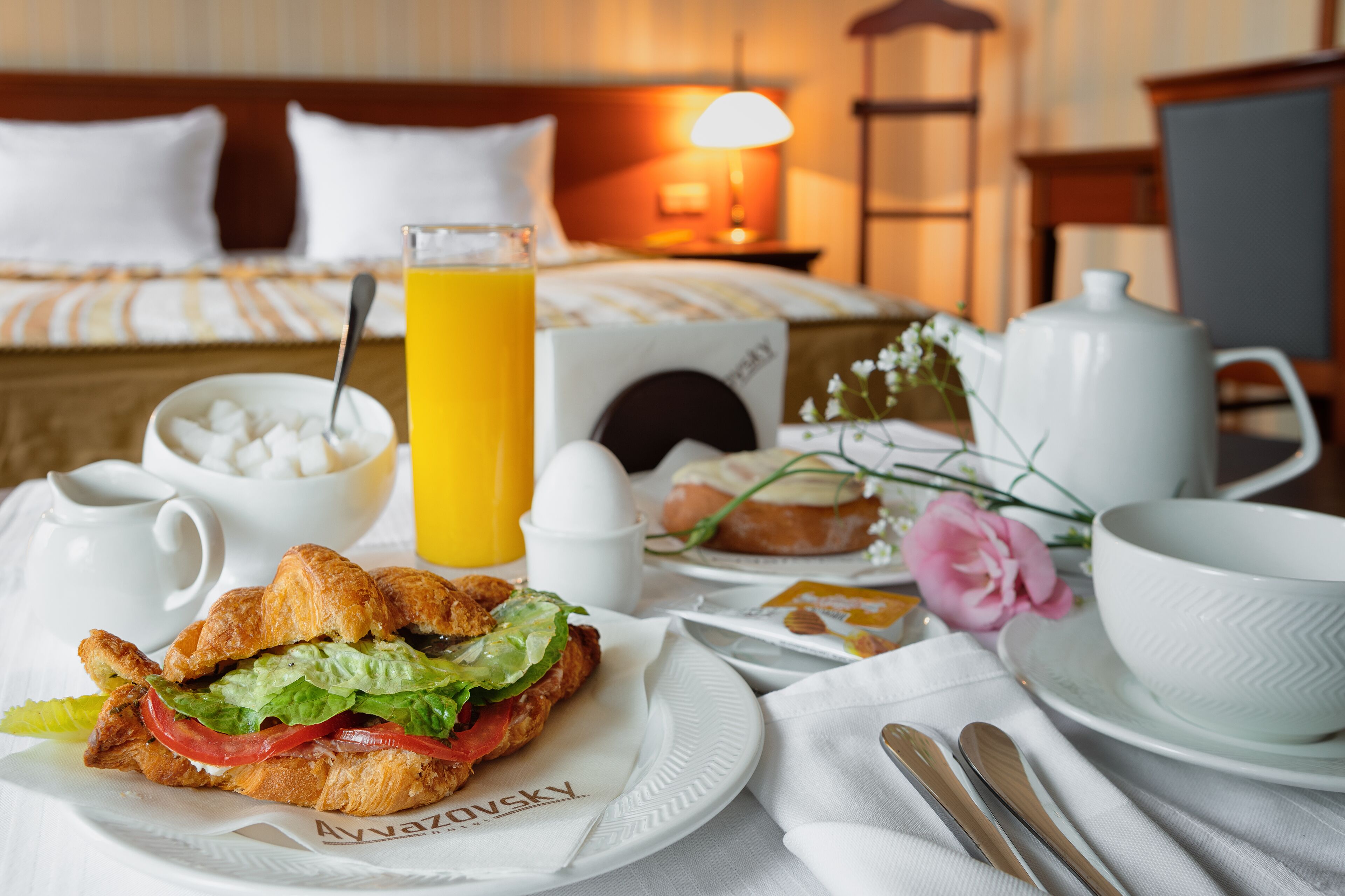 A breakfast setting with croissants, a sandwich, a glass of orange juice, a bowl of sugar cubes, a teapot, and a cup on a table.