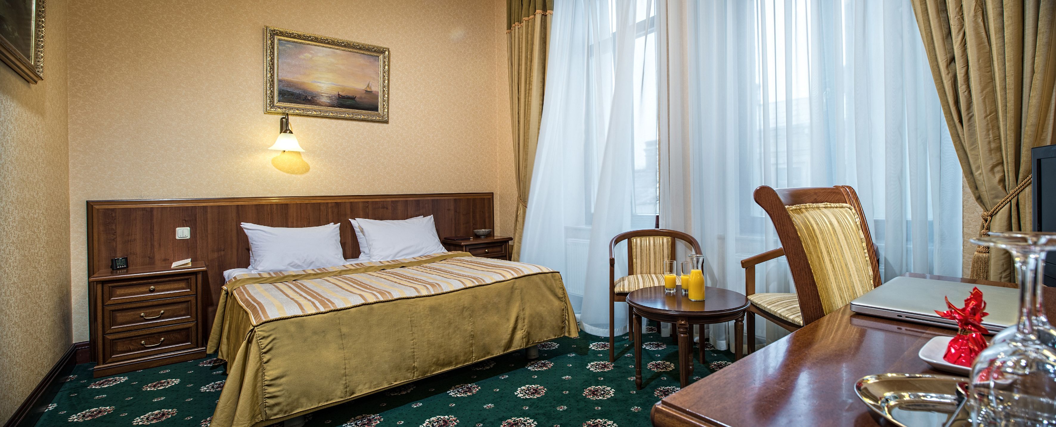 A hotel room with a bed, a desk with a laptop, a chair, a small table with glasses and a bottle, and a painting on the wall.