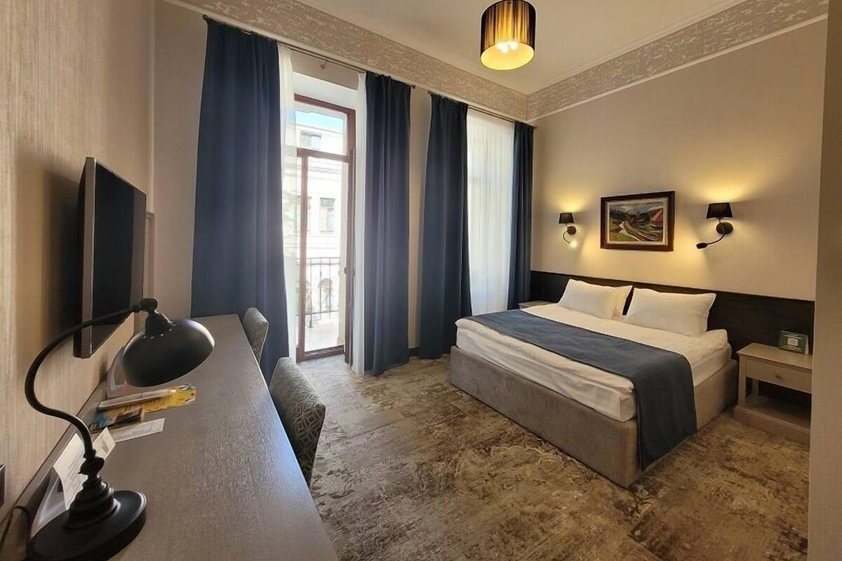 Et hotelværelse med seng, skrivebord med lampe, stol, et billede, vægmonteret lampe og udsigt ud af et vindue med gardiner.