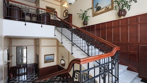 A grand staircase with a wooden handrail and black metal balusters, leading to a second floor. A large potted plant and framed artwork adorn the walls. - Ayvazovsky (Odesa)