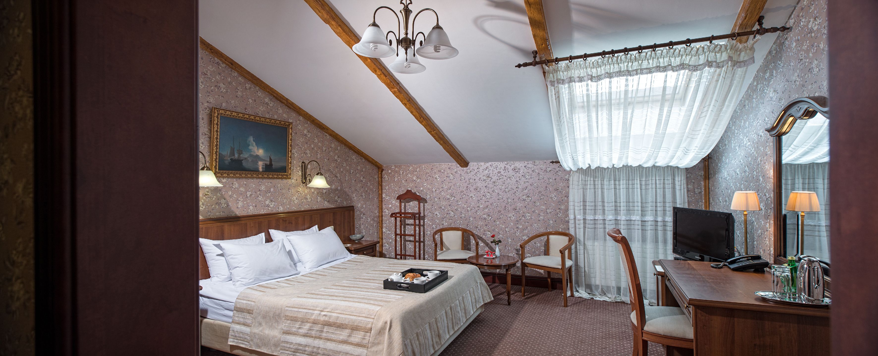 A hotel room with a bed, two chairs, a desk with a TV, a lamp, and a painting on the wall.