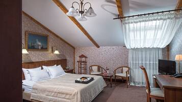 A hotel room with a bed, two chairs, a desk with a TV, a lamp, and a painting on the wall.
