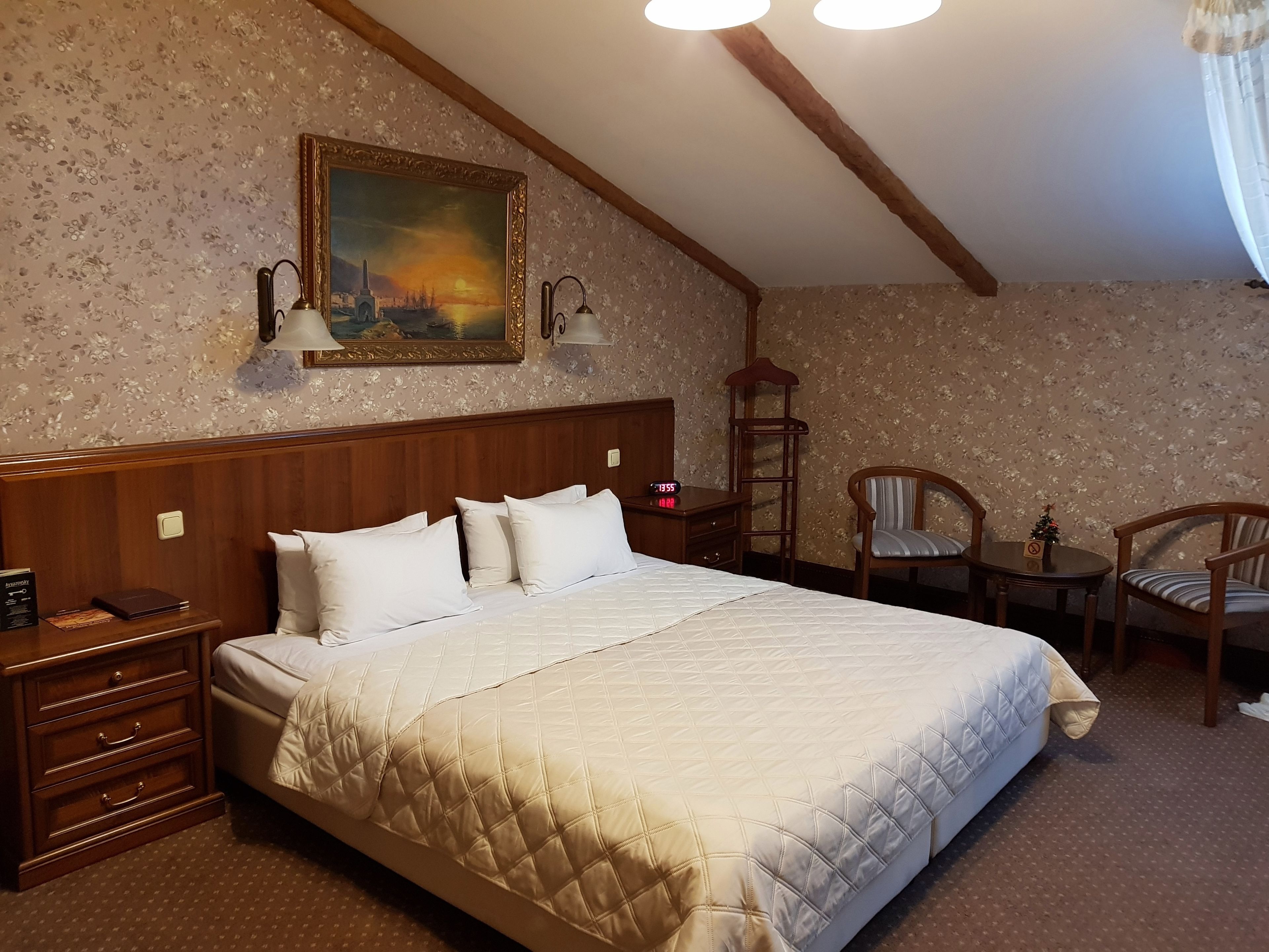 A hotel room with a large bed, wooden headboard, a nightstand, a desk with a chair, and a painting on the wall.