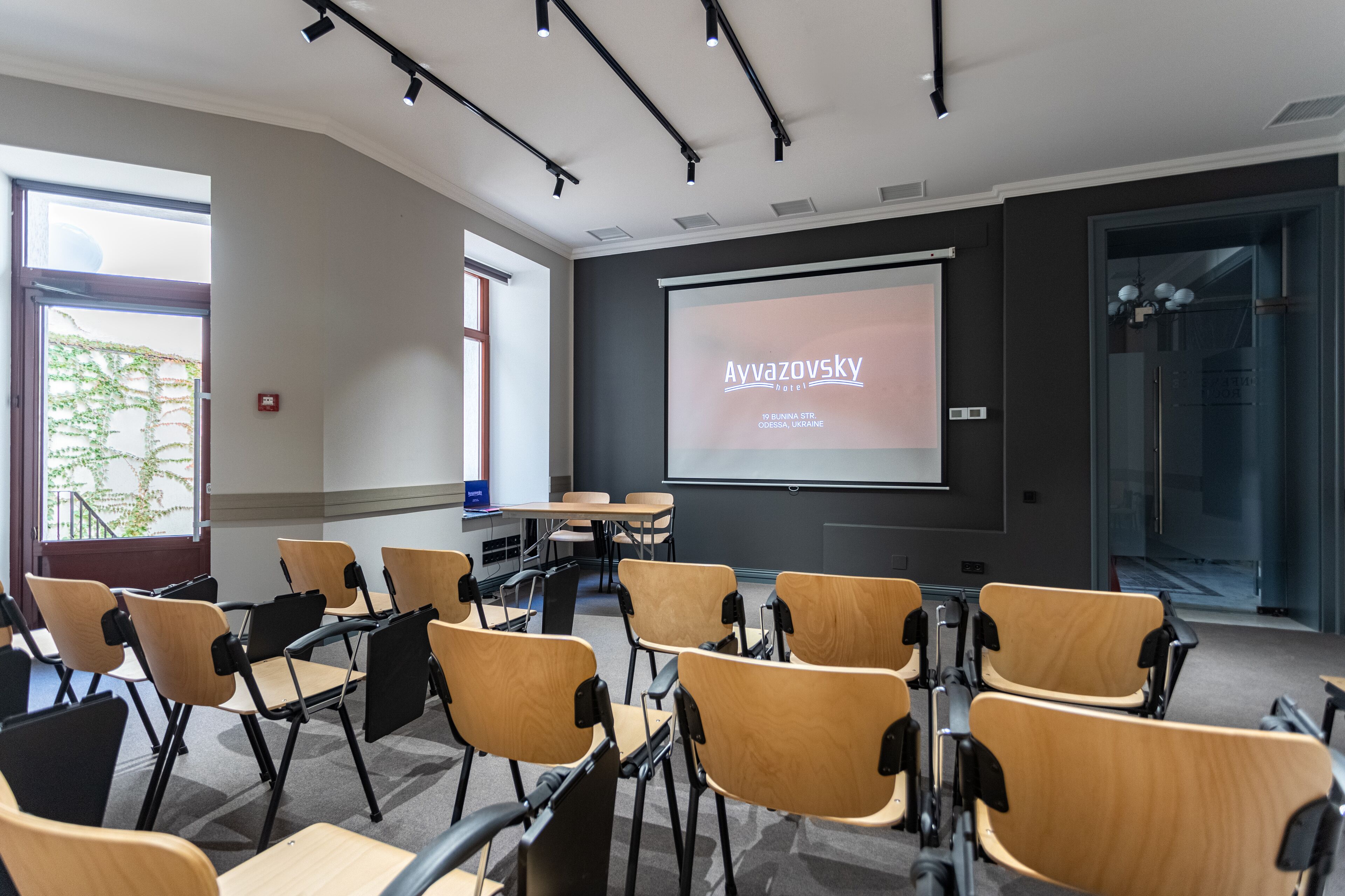 Un salón de conferencias con filas de sillas, una pantalla de proyector mostrando 'Avvaazovsky hotel' y un gran ventanal con vistas a zonas verdes.