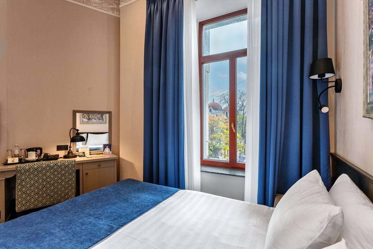 Et hotelværelse med en seng, et skrivebord med en computer, en lampe og et vindue med gardiner.