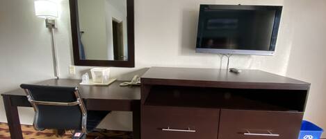 Una habitación de hotel con escritorio, silla, espejo, televisor y un bote de basura.