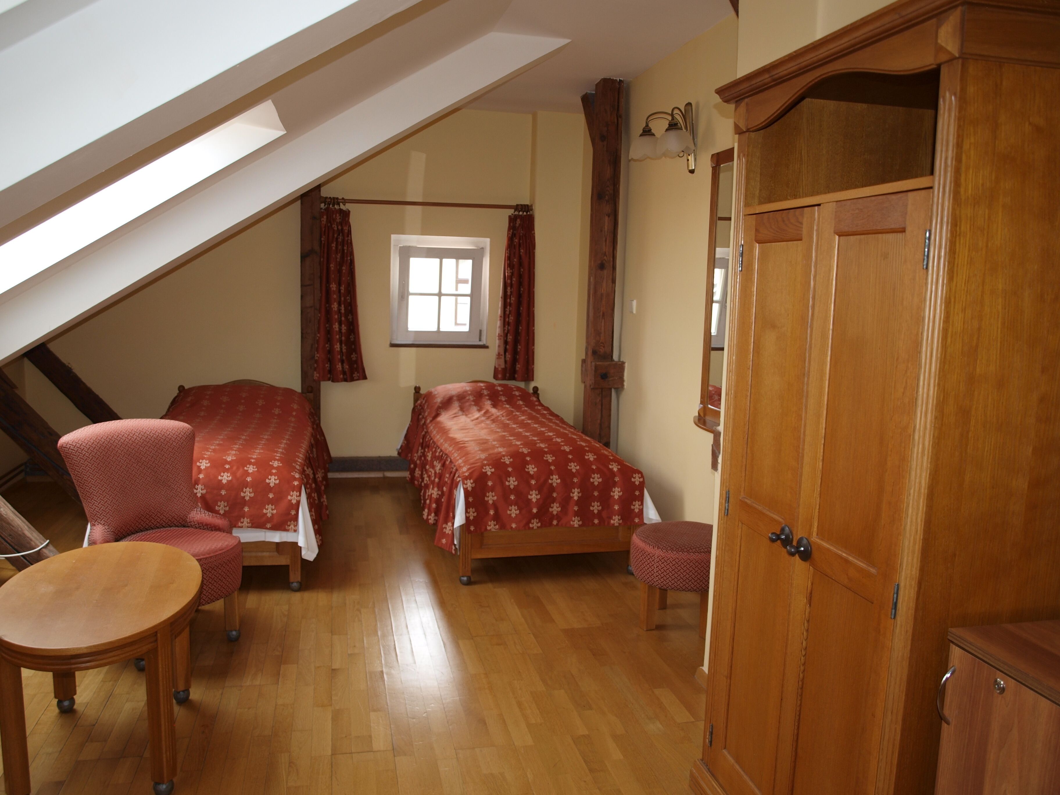 A bedroom with two beds, a wooden wardrobe, a small table, a chair, and a window with curtains.