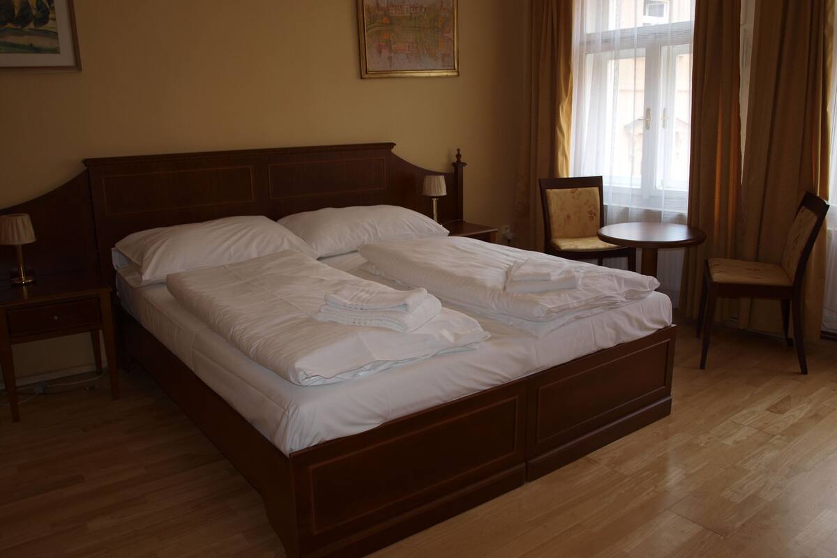 Un letto matrimoniale con lenzuola bianche, una testiera in legno, un comodino con una lampada, un tavolino con una sedia e un quadro appeso al muro.