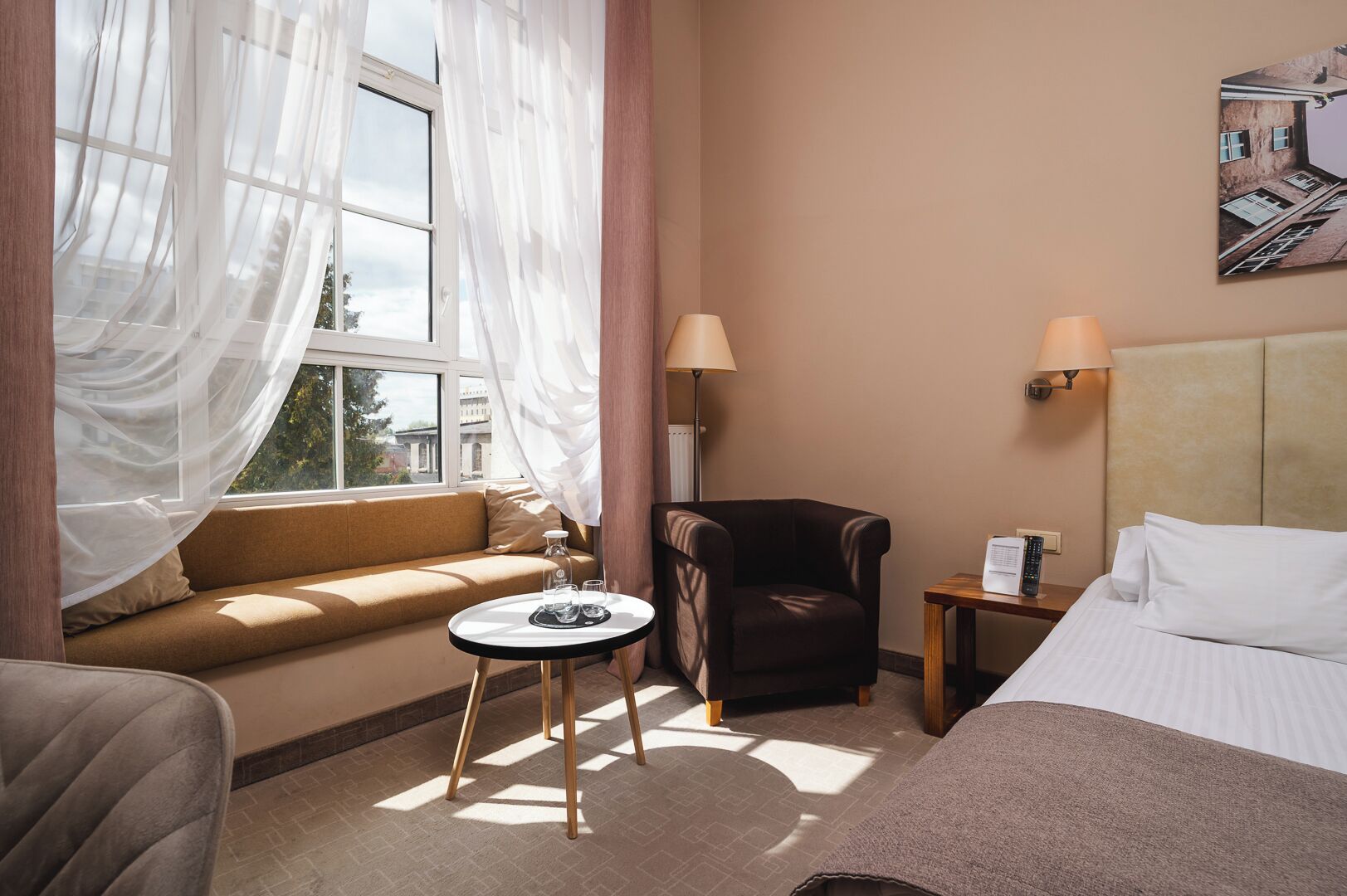 A hotel room with a sofa, a small table with glasses, a chair, a bed, and a wall-mounted lamp.