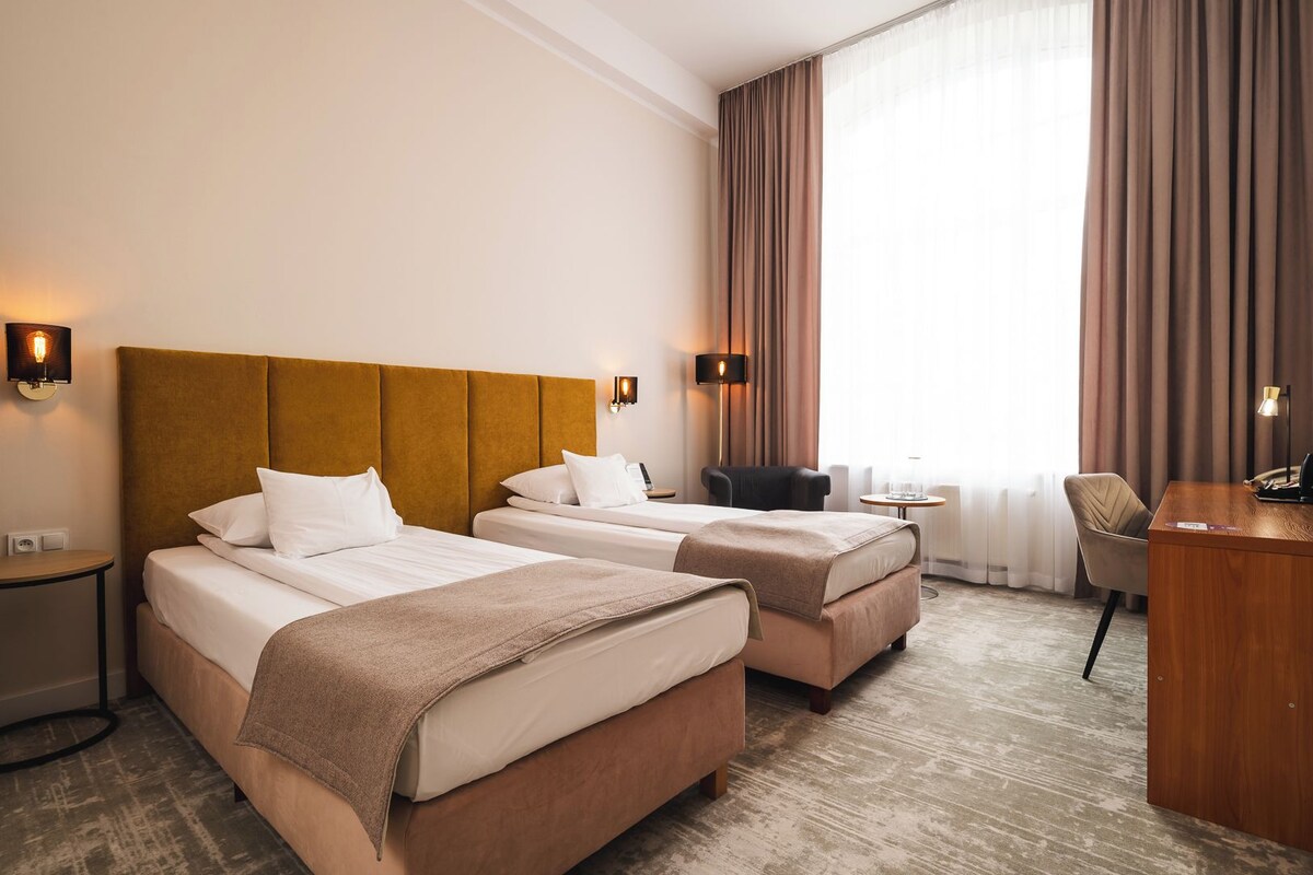 A hotel room with two beds, a desk, a chair, and a lamp.