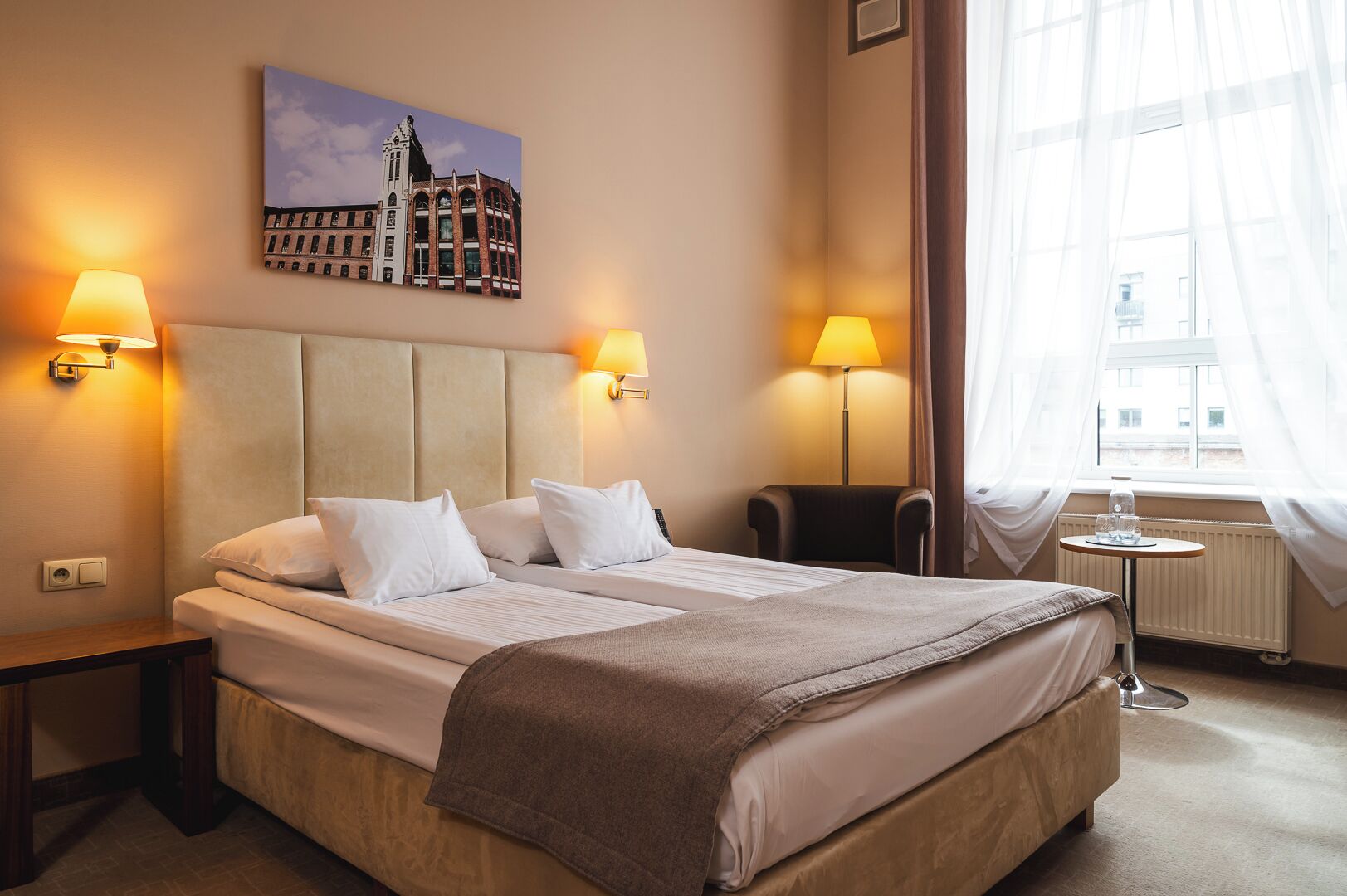 A hotel room with a large bed, two bedside lamps, a chair, a small table, and a window with sheer curtains.
