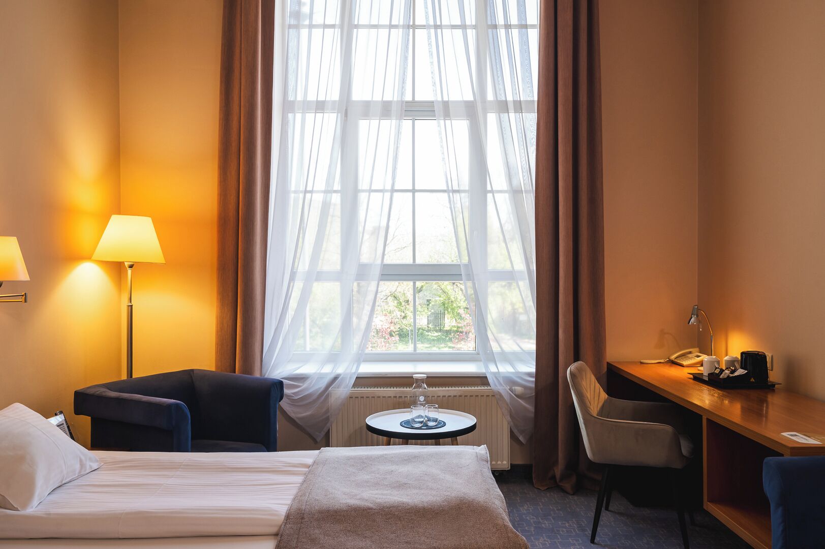 Habitación de hotel con cama, escritorio, silla, lámpara y vista por la ventana.