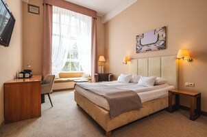 A hotel room with a bed, a desk, a chair, a window with curtains, a lamp, and a wall-mounted artwork.