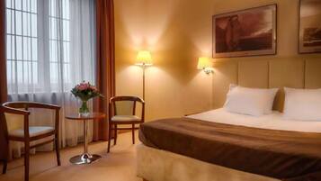 Una habitación de hotel con cama, dos sillones, una mesita de noche con un florero, dos lámparas de pared y un cuadro en la pared.