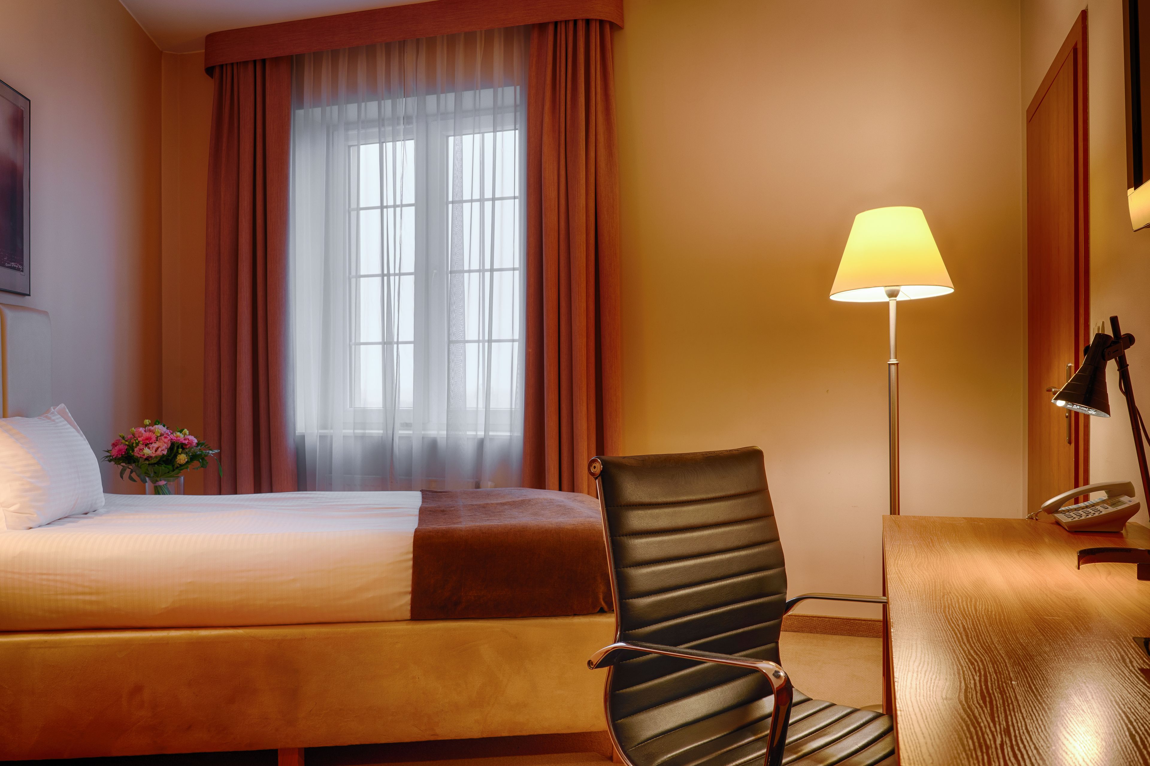 Una habitación de hotel con una cama, una silla, un escritorio con lámpara y un ventanal con cortinas.
