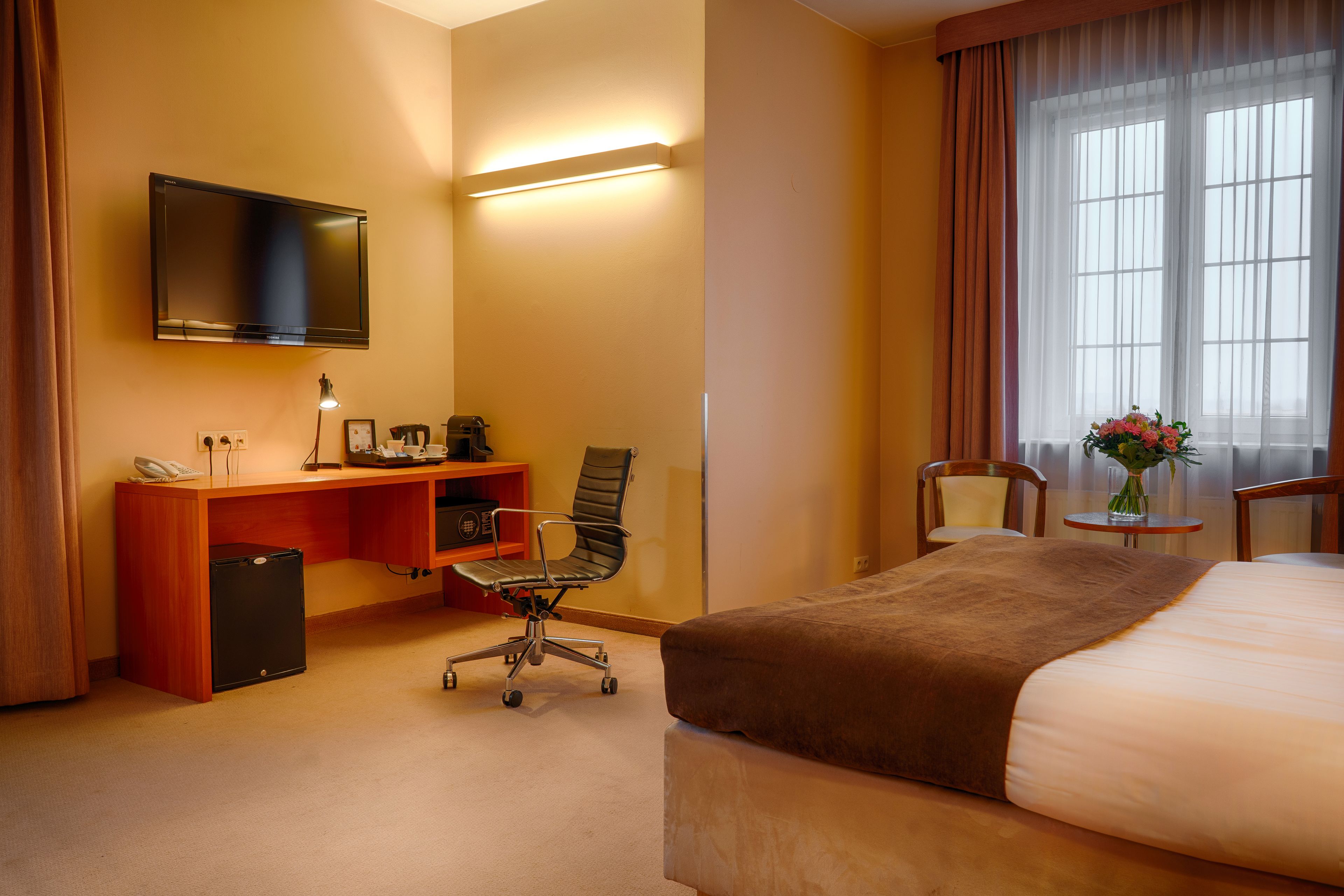 Ett hotellrum med en säng, ett skrivbord, en stol, en TV och ett litet bord med en vas.
