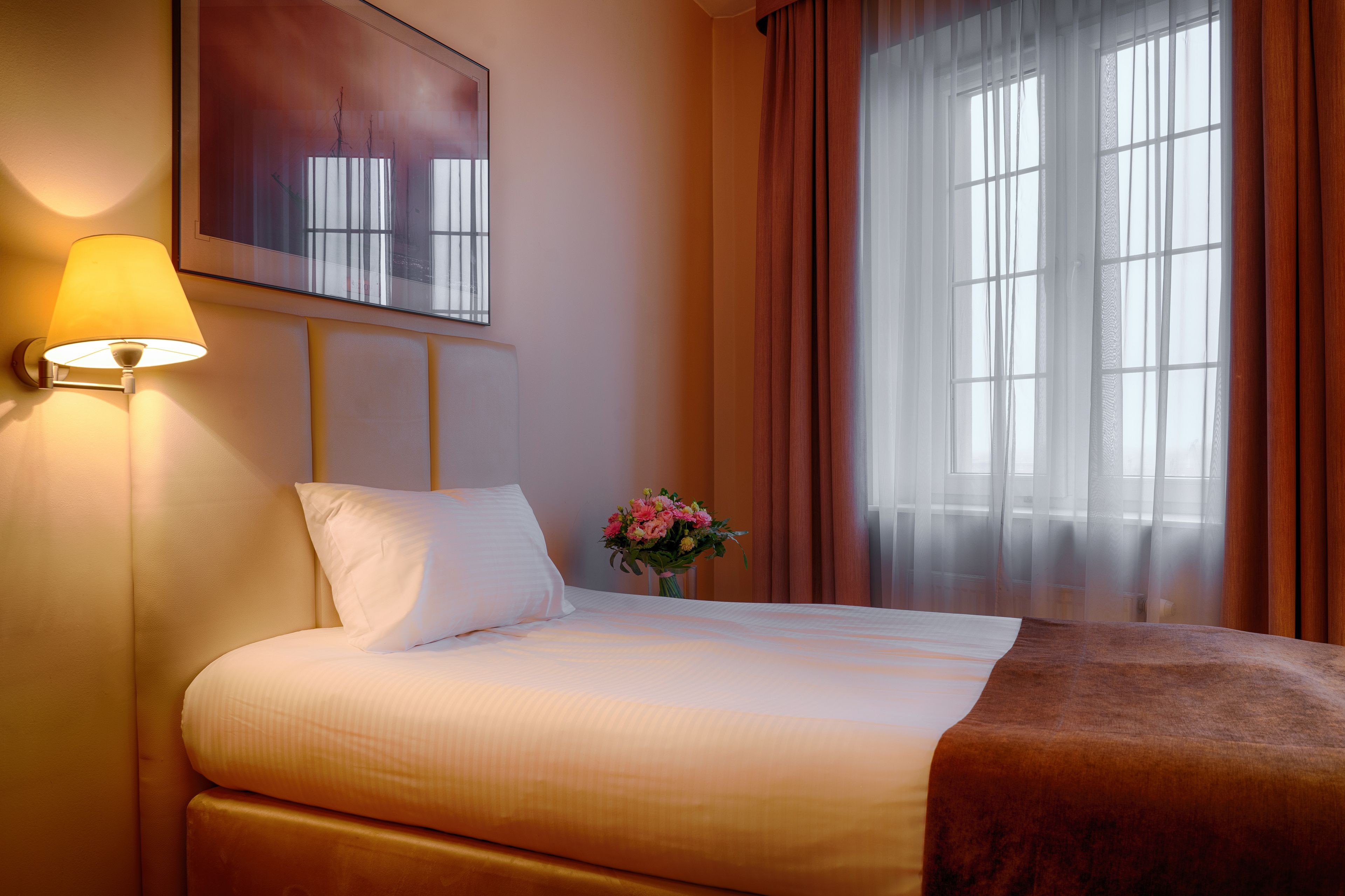 A hotel room with a bed, a lamp, a painting, a window with curtains, and a vase with flowers.