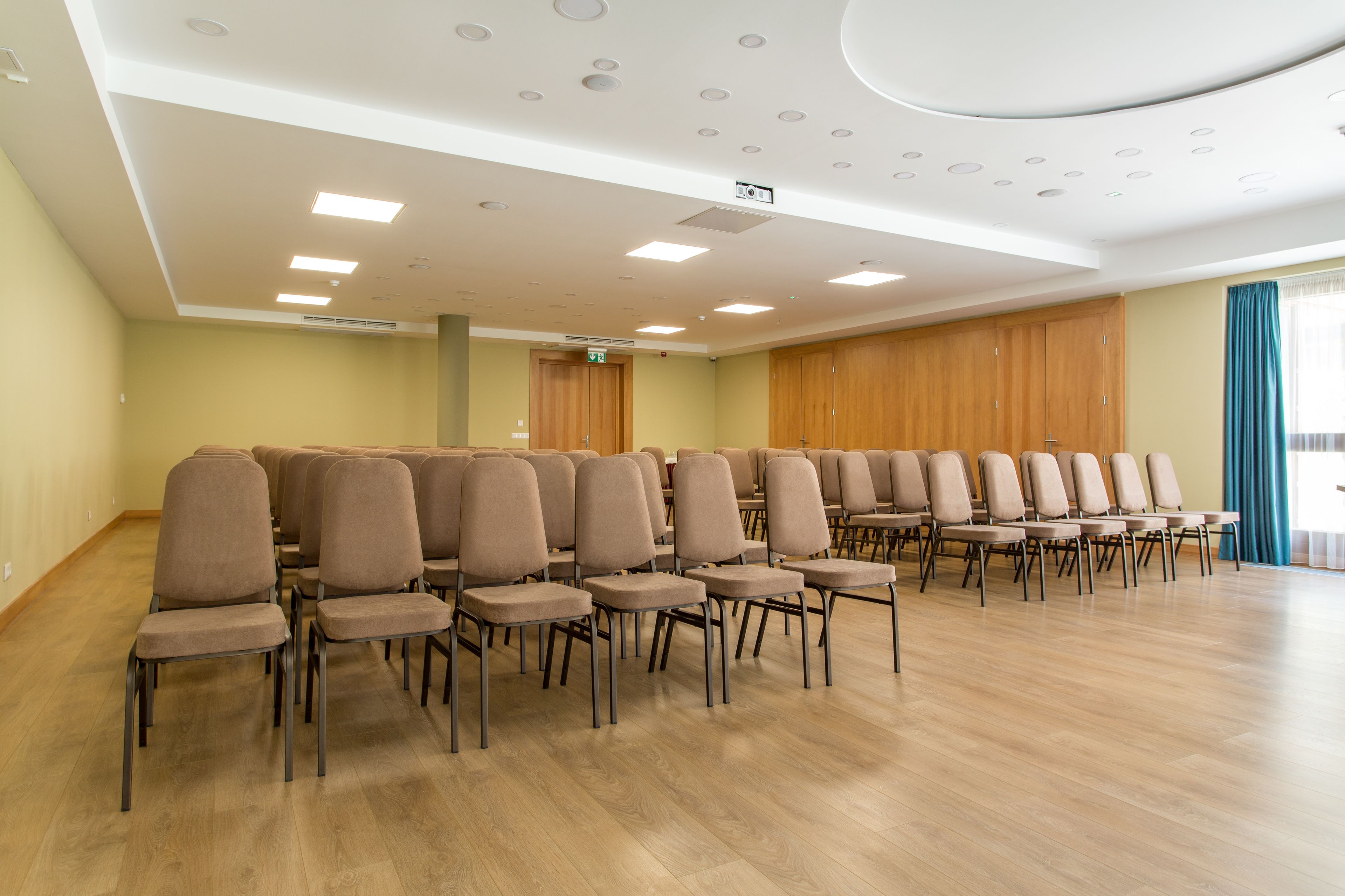 A conference room with rows of chairs, a wooden floor, and a white ceiling with recessed lighting.
