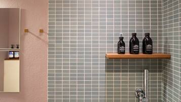 A bathroom with a tiled wall, a wooden shelf with three bottles, and a shower.