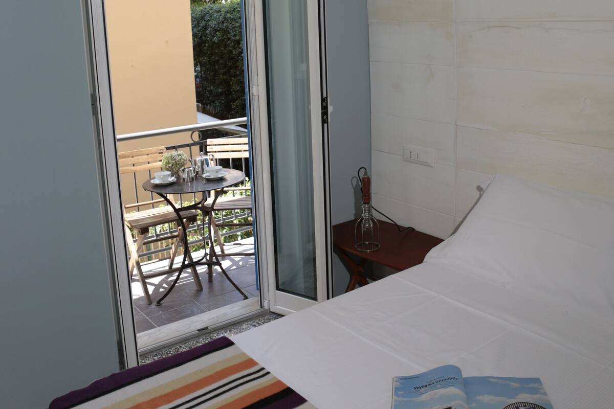 Een hotelkamer met een bed, een raam met uitzicht op een buitentafel en stoelen, en een tijdschrift op het bed.
