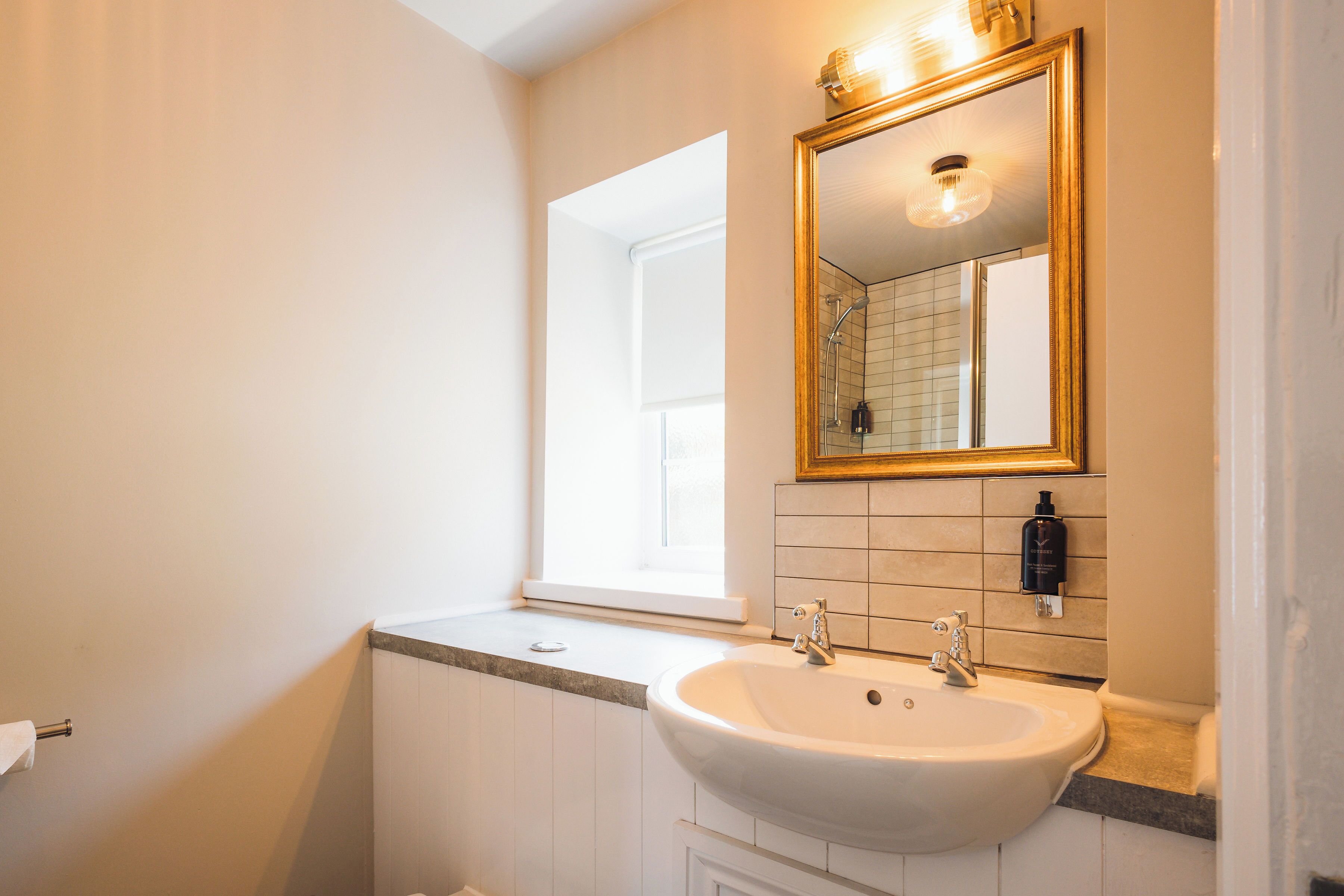 Un baño con un lavabo blanco, un espejo con marco dorado y una ventana.