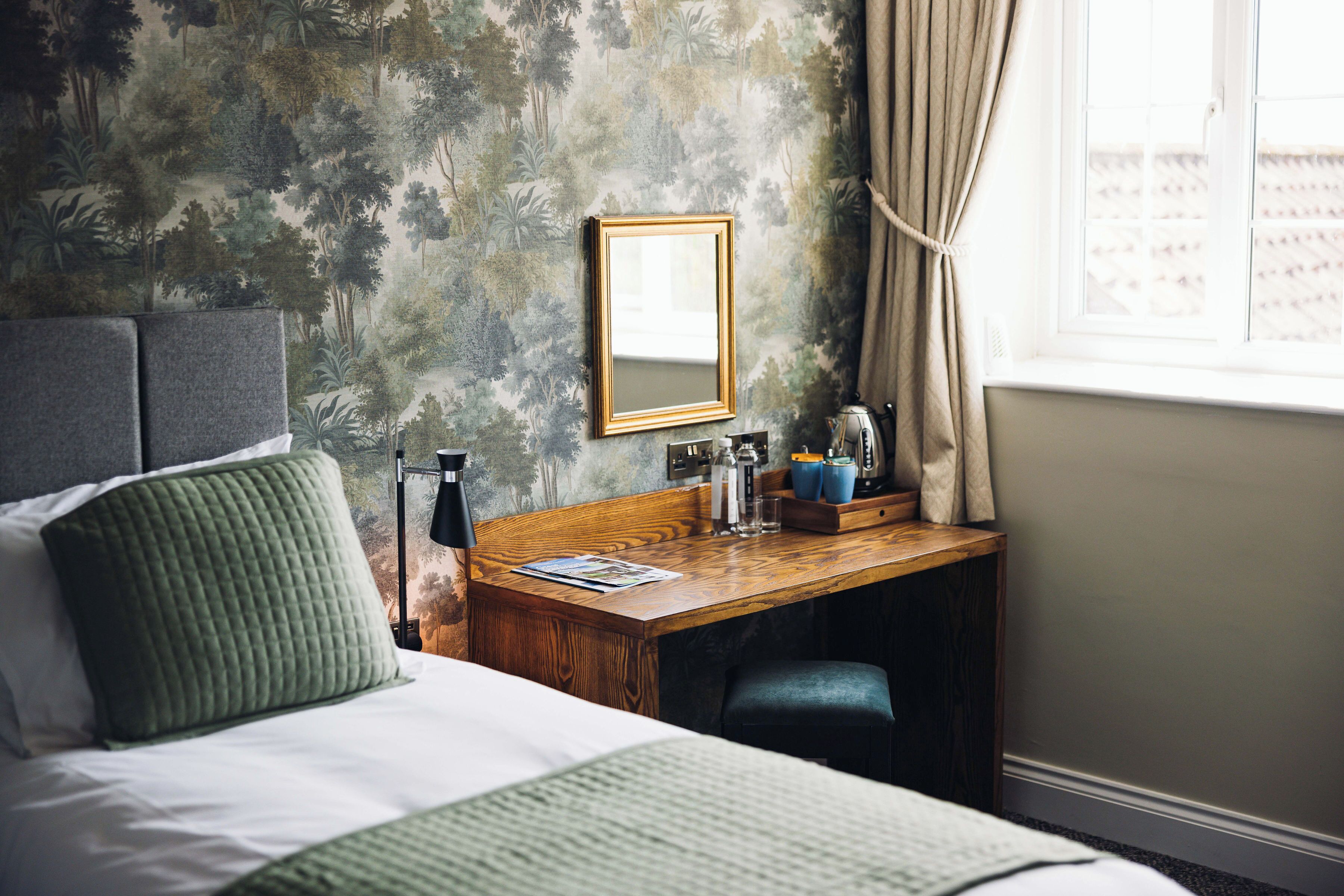 Une chambre d’hôtel avec un lit, un bureau doté d’un miroir, une fenêtre avec des rideaux et un mur orné d’un motif représentant une forêt.