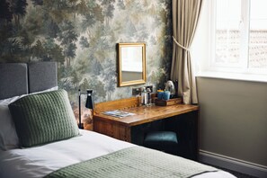 A hotel room with a bed, a desk with a mirror, a window with curtains, and a wall with a forest pattern. - The George and Dragon Hotel (York)