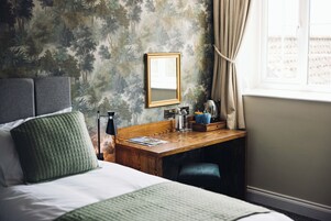 A hotel room with a bed, a desk with a mirror, a window with curtains, and a wall with a forest pattern.