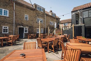 An outdoor patio area with multiple wooden tables and chairs, stone buildings in the background, and a clear sky. - The George and Dragon Hotel (York)