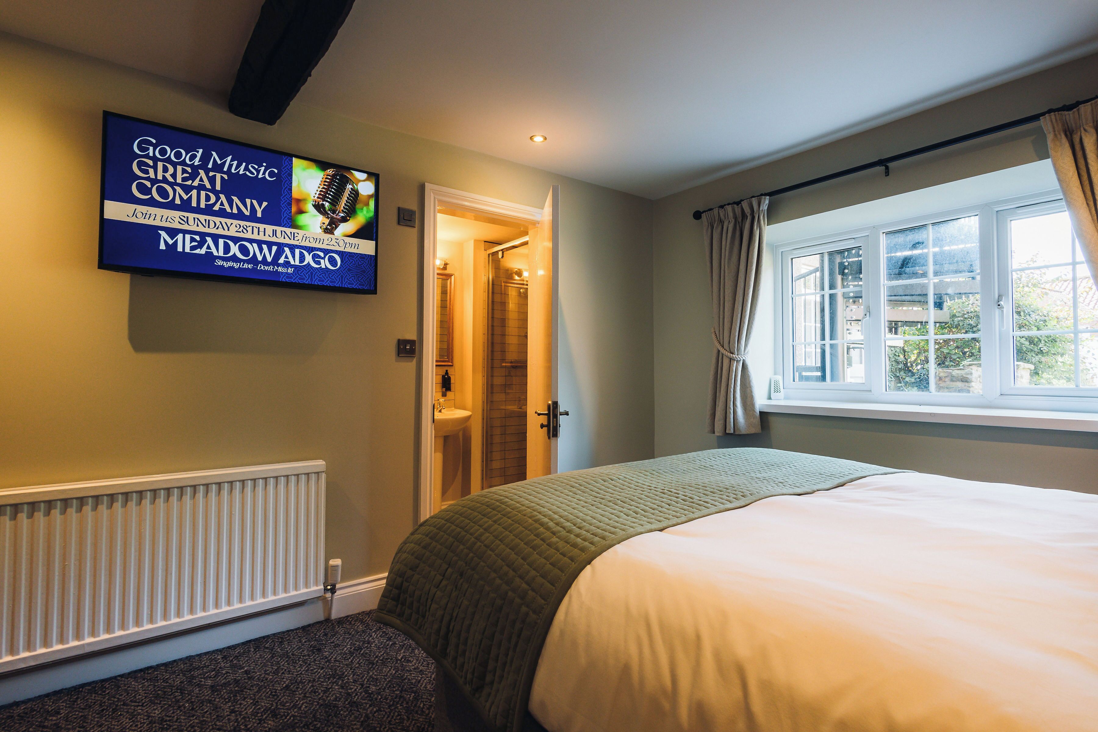 A bedroom with a bed, a radiator, a television displaying an advertisement, a window with curtains, and a door leading to a bathroom.