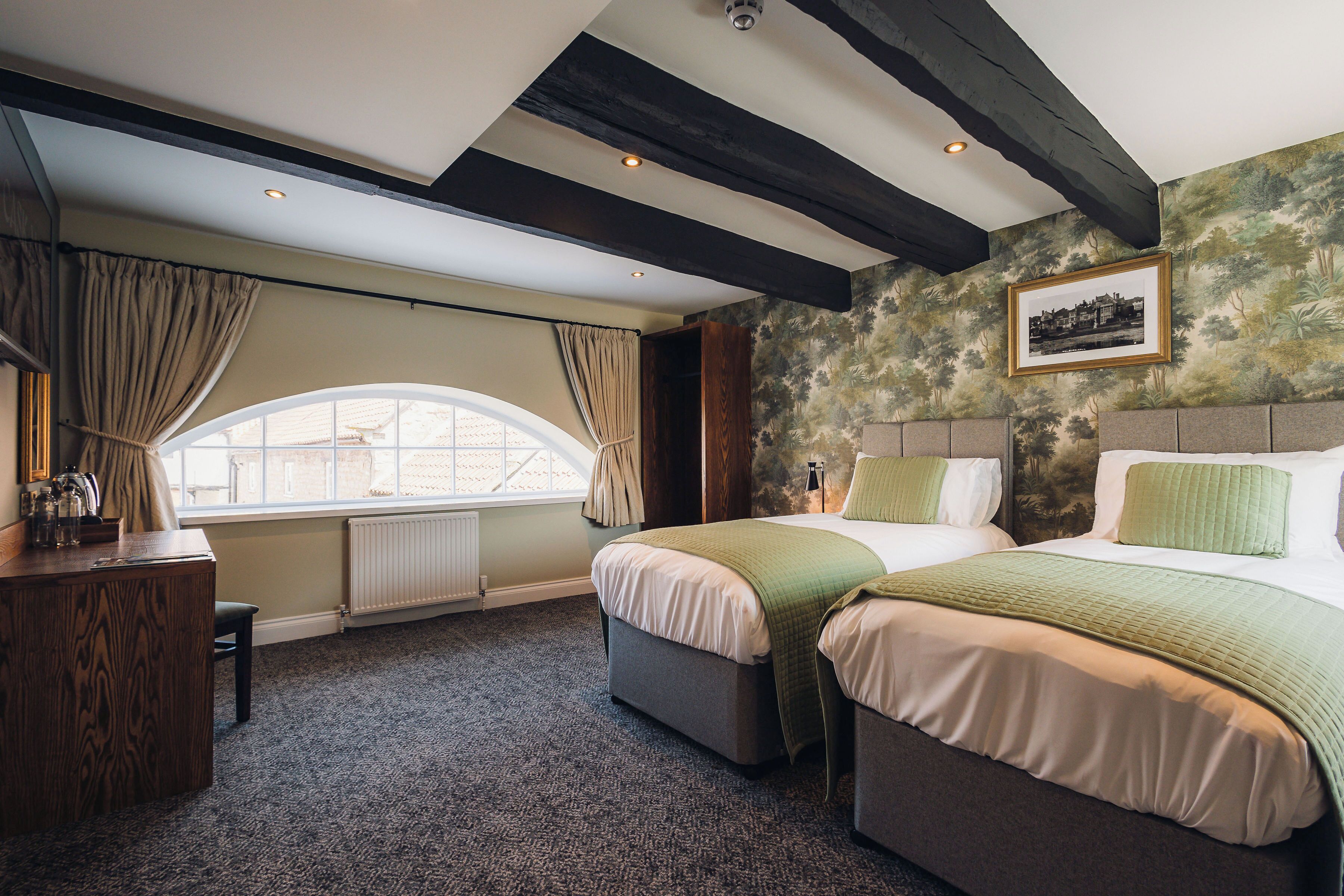 A hotel room with two beds, a desk, a window with curtains, and a fireplace.