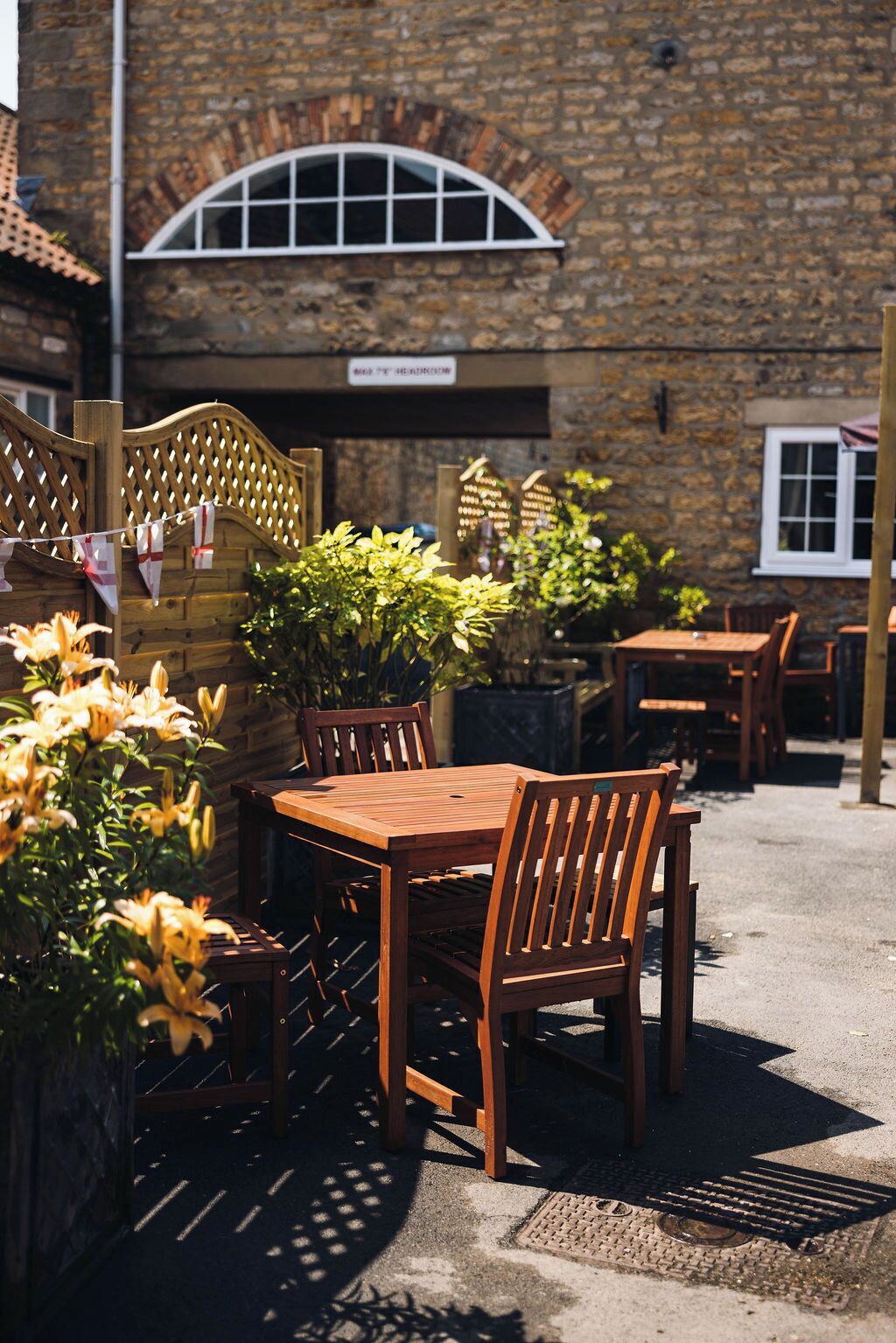 An outdoor wooden table and chairs set against a stone building with a brick arch window.