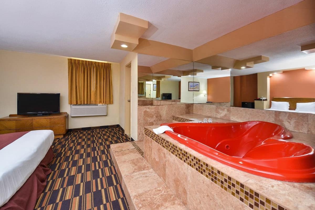 A hotel room with a large bed, a red jacuzzi, a television, and a mirror.