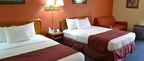 A hotel room with two beds, a nightstand, a lamp, a chair, and a small table.