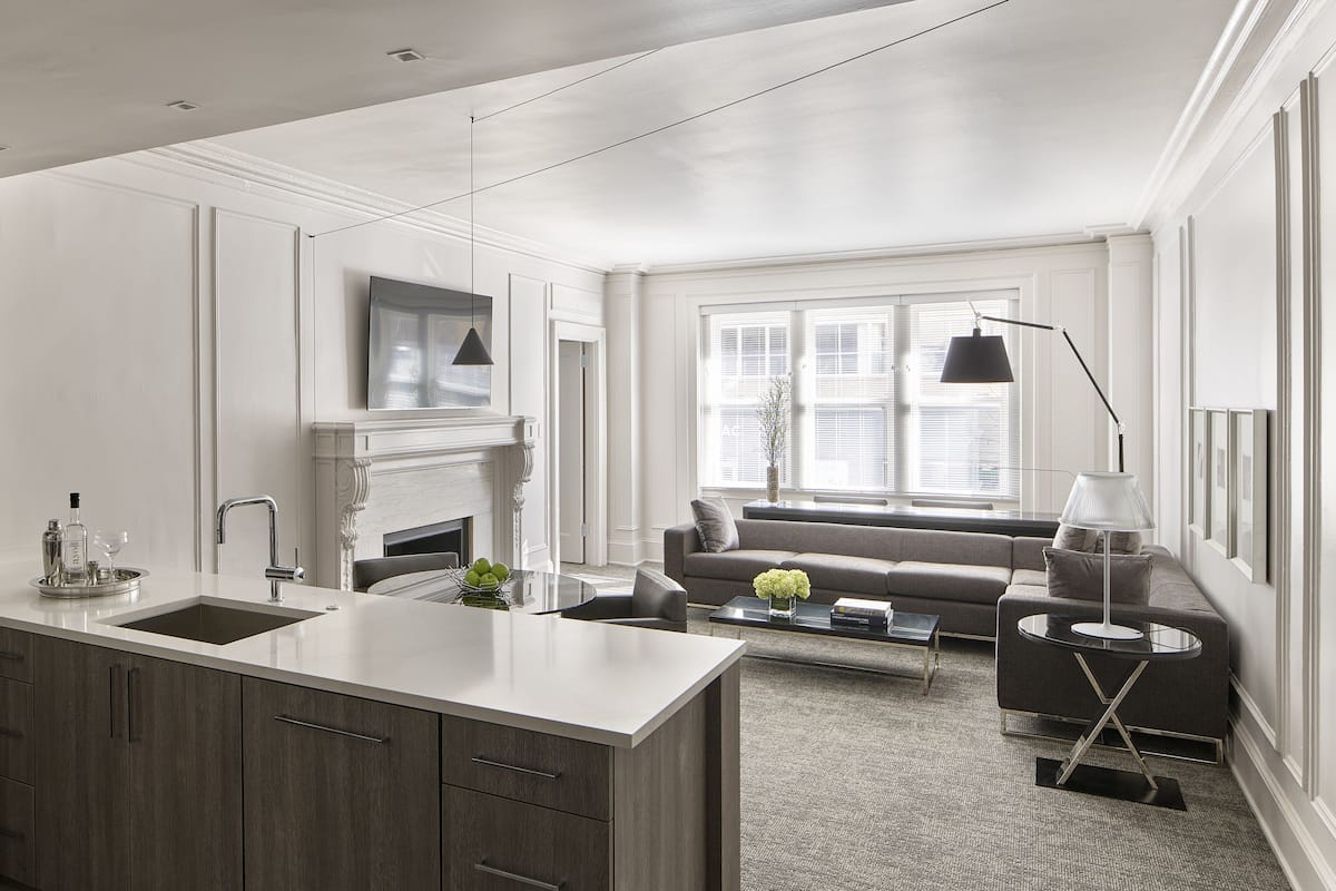 A modern kitchen with a central island, a dining area with a sofa and coffee table, and a living room with a fireplace and large windows.